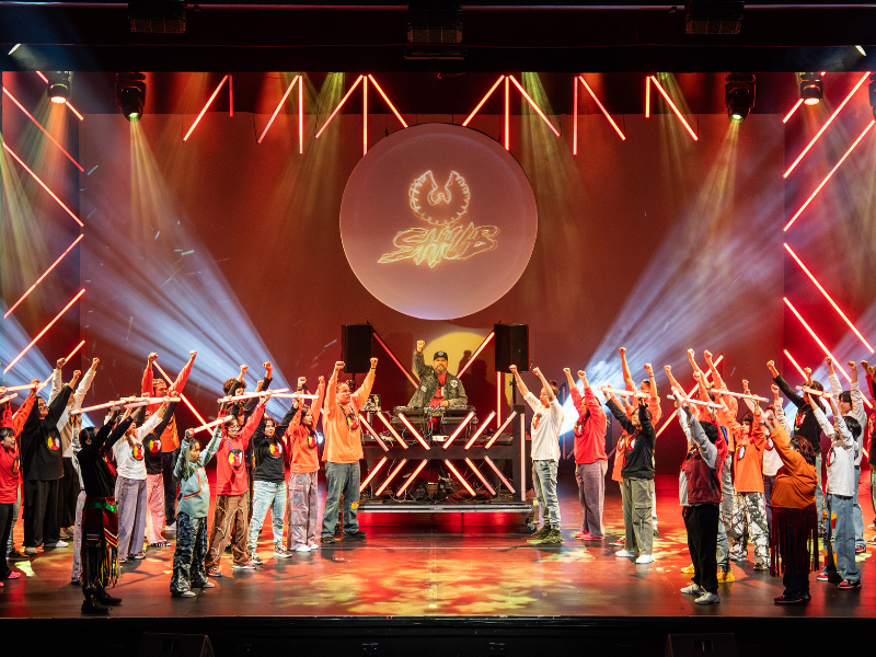 A large number of dancers are on the stage with light beams surrounding them. They are all dressed in similar attire. 